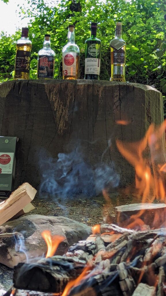 Man sieht ein loderndes Feuer und darüber die auf einem Baumstumpf thronenden fünf Flaschen Whisky aus dem nächsten Bild.